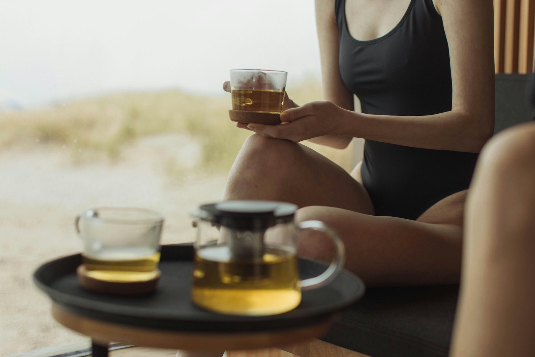 Person sitting by a body of water holding a glass of tea with a tray of tea on a small table.